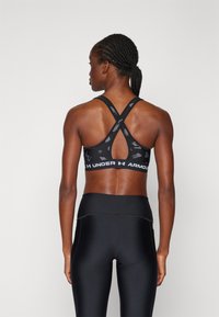 Melna sporta krūšturis ar krustveida siksnu dizainu un teksturētu rakstu. Iezīmēts ar vienkāršu joslu, kurā ir "Under Armour" zīmols.