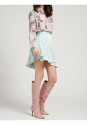 Kvinde iført blomstret bluse med lange ærmer, lyseblå nederdel med polkaprikker og flæser samt lyserøde knæhøje støvler med hæl, stående foran ensfarvet baggrund.