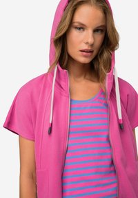 Giacca con cappuccio e zip in tessuto rosa brillante con tasca frontale, indossata sopra una maglietta a righe blu e rosa, dotata di maniche corte e dettagli con cordini.