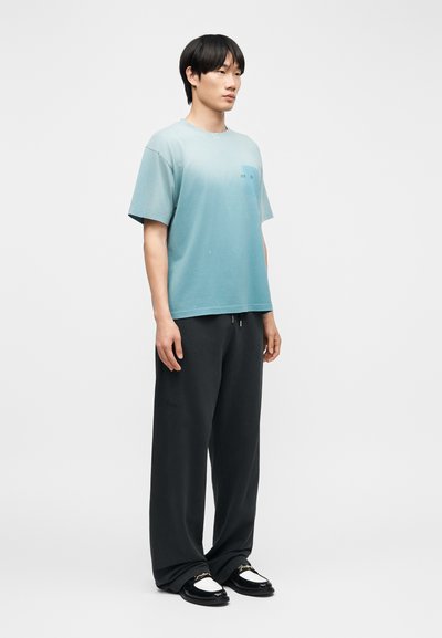 Νέος άνδρας που στέκεται, φοράει ανοιχτό μπλε φαρδιά μπλούζα, φαρδιά μαύρη παντελόνια και μαύρα-άσπρα μοκασίνια με λεπτομέρεια αλυσίδας.