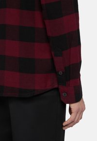Camisa de franela a cuadros rojos y negros con puños de botón, que presenta una abertura lateral y una textura suave. El tejido parece cálido y resistente.