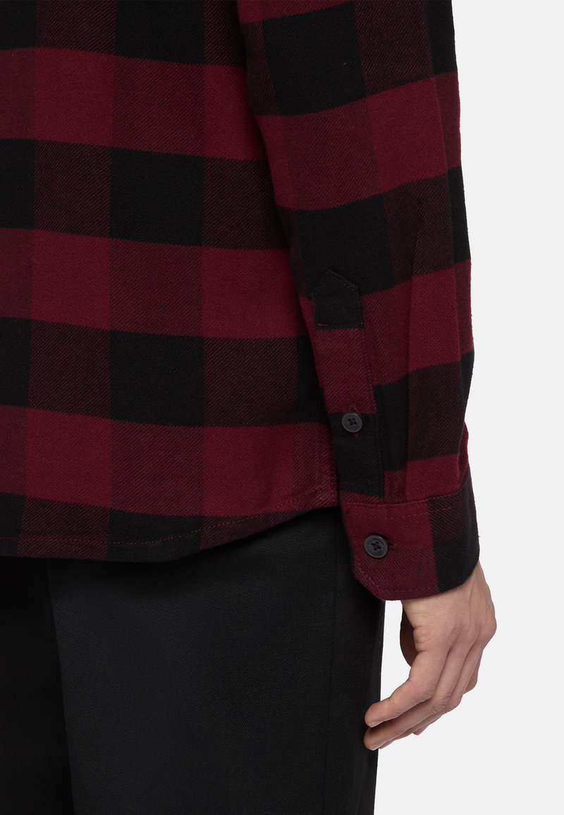 Camicia in flanella a quadretti rossi e neri con polsini a bottone, dotata di una fessura laterale e una texture liscia. Il tessuto appare caldo e resistente.