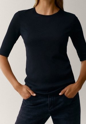 Vrouw draagt een strakke, zwart geribbelde T-shirt met korte mouwen, met handen in de zakken, zichtbaar van nek tot heupen tegen een effen achtergrond.