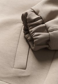 Beige sweatshirt kain dengan cuff yang dikumpulkan, tekstur halus, dan detail jahitan yang halus pada lengan dan badan.