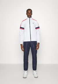 Hvit jakke med røde og marineblå striper, glidelås foran, kombinert med marineblå bukser med et logo. Brukt med hvite sneakers.