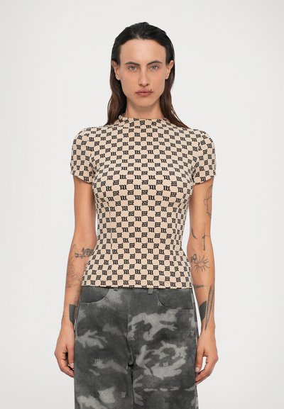 Donna con lunghi capelli scuri che indossa una maglietta beige aderente con un motivo nero e pantaloni mimetici grigi, in piedi su uno sfondo semplice.