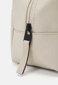 Beige, teksturert håndveske med en svart glidelåsdrager og lys beige stropp. Har synlige sømmer og en glatt, matt finish.