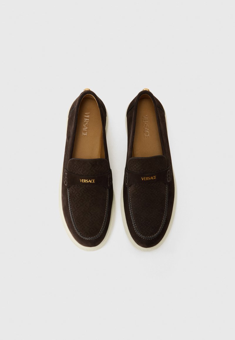 Braune Wildleder-Slipper mit einem strukturierten Muster, versehen mit einem goldenen "Versace"-Logo auf einem Riemen und einer kontrastierenden weißen Sohle.