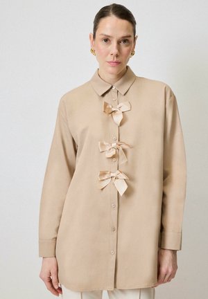 Beige button-up shirt with a collar features three ribbon bows as accents down the front. Long sleeves with buttoned cuffs. Smooth fabric texture.