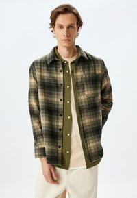 Chemise à carreaux verte et beige avec un col, superposée sur une chemise vert foncé. Un sous-vêtement beige clair est visible ; le mannequin se tient devant un fond uni.