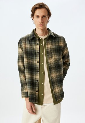 Camisa de cuadros verde y marrón claro con cuello, superpuesta sobre una camiseta verde oscuro. Camiseta interior beige clara visible; el modelo está de pie contra un fondo liso.