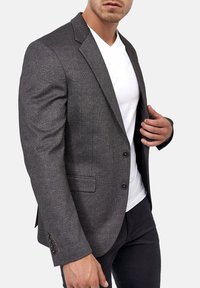 Blazer gris a medida con una tela texturizada, solapas con muesca, dos botones y sutilezas en la costura. Llevado sobre una camisa blanca.