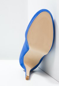 Modrá semišová lodička na vysokém podpatku, s špičatou špičkou a hladkou béžovou podrážkou. Podpatek je štíhlý a má texturovaný povrch.