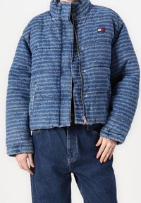 Blå puffjacka med ett mönster av text över hela plagget, hög krage och dragkedjestängning. Inkluderar en liten logotyp på vänster bröst.