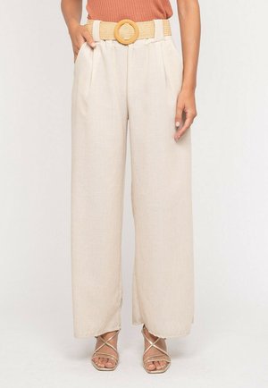 Pantalon beige à jambes larges avec des plis à l'avant, associé à une ceinture tissée à boucle ronde et des sandales dorées à brides sur une personne debout.