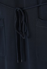Gros plan d’un pantalon bleu marine foncé avec un cordon noué à la taille et deux poches plaquées à l’avant.