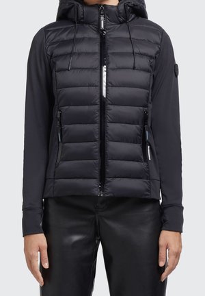 Czarna pikowana kurtka na zamek z kapturem i długimi rękawami, noszona z czarnymi spodniami, widok z przodu od talii do szyi.