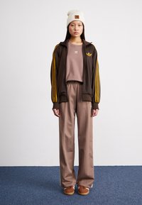 Brun joggebomulljakke med gule striper, beige crew neck-genser og løse brune bukser. Hvit beanie-lue og brune joggesko.