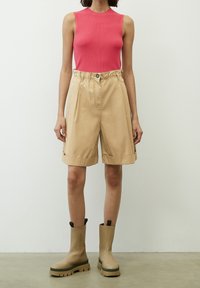 Persona che indossa una maglietta senza maniche rosa, pantaloni corti beige a vita alta fino al ginocchio con risvolti e stivali chunky beige, in piedi contro un muro bianco.