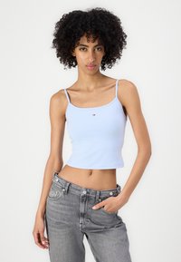 Noor naine lokkis juustega, seljas helesinine lühike T-särk ja hallid teksapüksid, seisab ühe käega taskus valge tausta vastu.