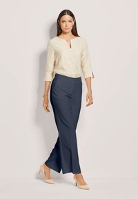 Vrouw in een gestructureerde crèmekleurige blouse met 3/4 mouwen, marineblauwe, op maat gemaakte wijde broek, beige hakken, die tegen een neutrale achtergrond loopt.
