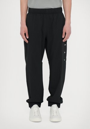 Hombre vistiendo pantalones jogger negros con cintura elástica y letras verticales blancas en la pierna izquierda, y zapatillas blancas, de pie contra un fondo liso.