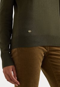 Oliven grøn genser med ribbet nederkant og mansjetter, med en liten metalllogo-detall. Båret over brune kordfløyelsbukser med en teksturert overflate.