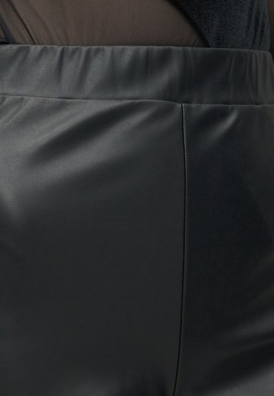 Close-up van de tailleband van een zwarte nep leren rok met stikseldetails, gedragen met een donkere doorschijnende top die erin is gestopt.