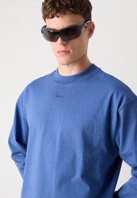 Blue long-sleeve t-shirt with a crew neckline, featuring a small logo near the collar. The model wears sleek black sunglasses.