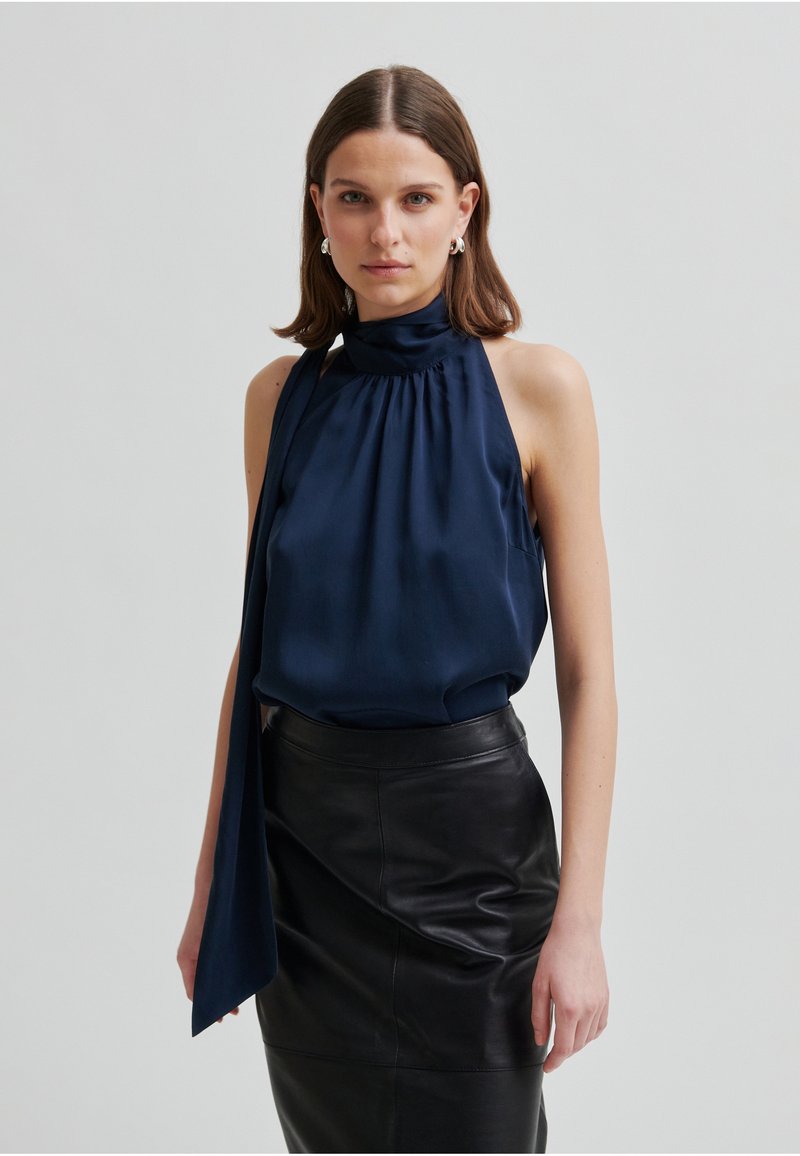 Marineblauw satijnen haltertop met een hoge hals en strikdetail, gecombineerd met een nauwsluitende zwarte leren rok met een gladde textuur.