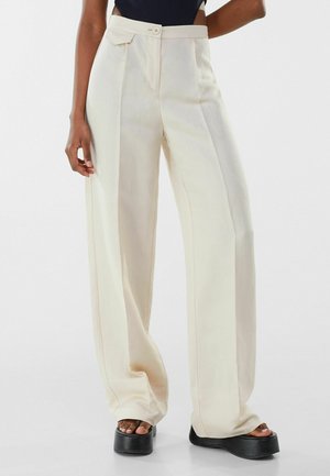 Vrouw draagt een crème broek met hoge taille en wijde pijpen met plooien aan de voorkant en zwarte platformsandalen, staand tegen een effen achtergrond.