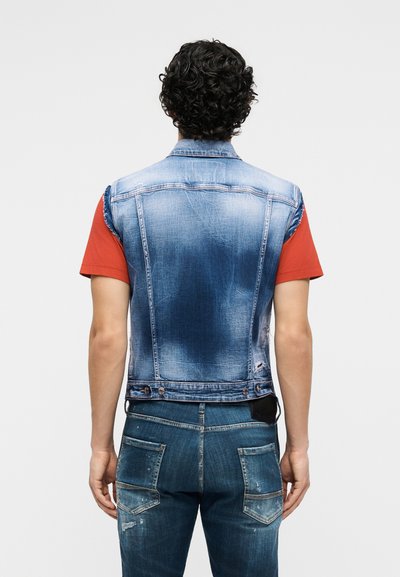 Άνδρας με σγουρά μαύρα μαλλιά που φοράει ξεθωριασμένο μπλε αμάνικο τζιν μπουφάν πάνω από κόκκινο κοντομάνικο πουκάμισο και σκισμένο μπλε τζιν, με την πλάτη γυρισμένη.