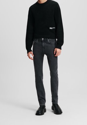 Person trägt einen schwarzen Langarmpullover mit kleinem weißem Logo, ausgewaschene schwarze Slim-Fit-Jeans und schwarze Lederstiefel auf neutralem Hintergrund.