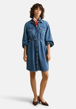 Femme debout portant une robe en jean longueur genou avec des manches retroussées, une ceinture fine, un foulard coloré, et des escarpins marron à bouts pointus.