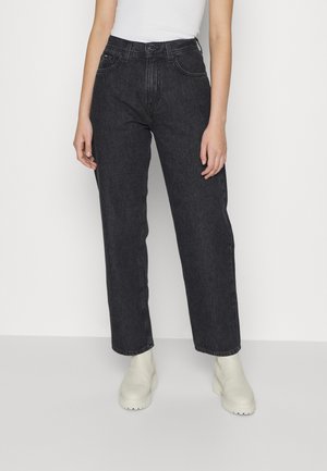 Person iført sorte jeans med højtaljede lige ben, hvid ærmeløs top og lyse ankelstøvler, stående mod en ensfarvet baggrund.