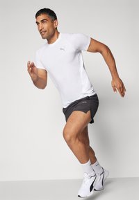 Valge sportlik T-särk lühikeste varrukatega ja Puma logo; kombineeritud tumedate lühikeste püksidega ja valgete tossudega mustade aktsentidega. Jooksupositsioon.