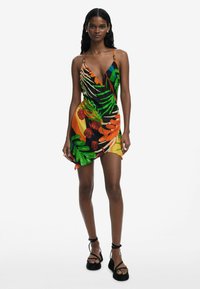 Vestido de estampado tropical con diseño envolvente, que presenta vibrantes tonos verdes, naranjas y rojos sobre un fondo negro. Tirantes finos y dobladillo asimétrico.
