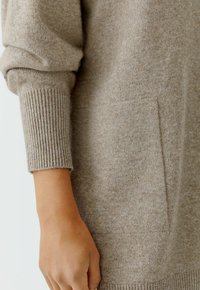 Beiger Strickpullover mit weicher Textur, ausgestattet mit gerippten Bündchen und einer dezenten Seitentasche. Nahaufnahme, die die Stoffdetails und die Armposition zeigt.