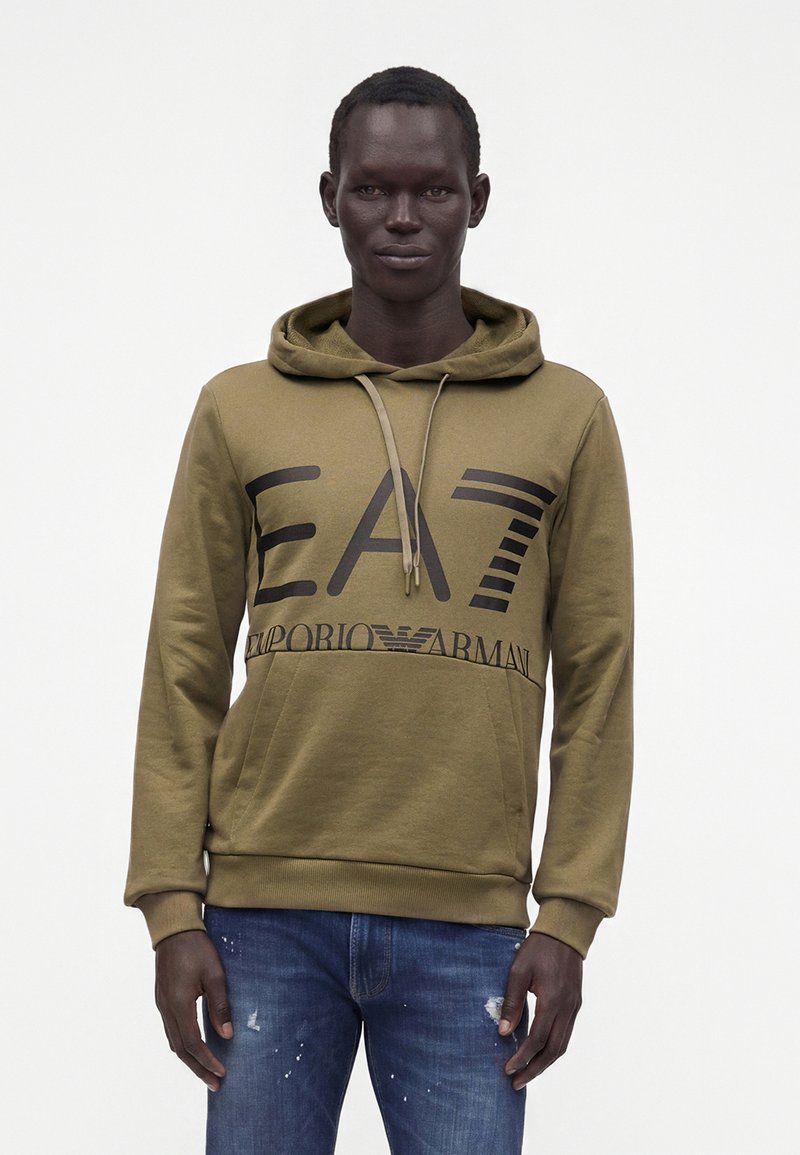 Άνδρας που φοράει χακί φούτερ Emporio Armani EA7 και ξεφτισμένο μπλε τζιν, στέκεται μπροστά σε απλό λευκό φόντο.
