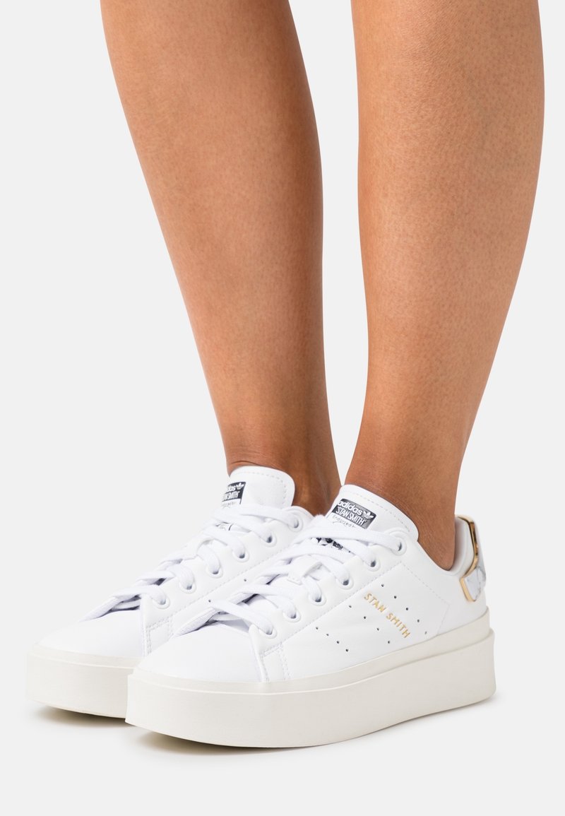 Sneakers bianche basse in pelle liscia, con lacci piatti, perforazioni circolari e suola platform rialzata, caratterizzate da dettagli dorati.