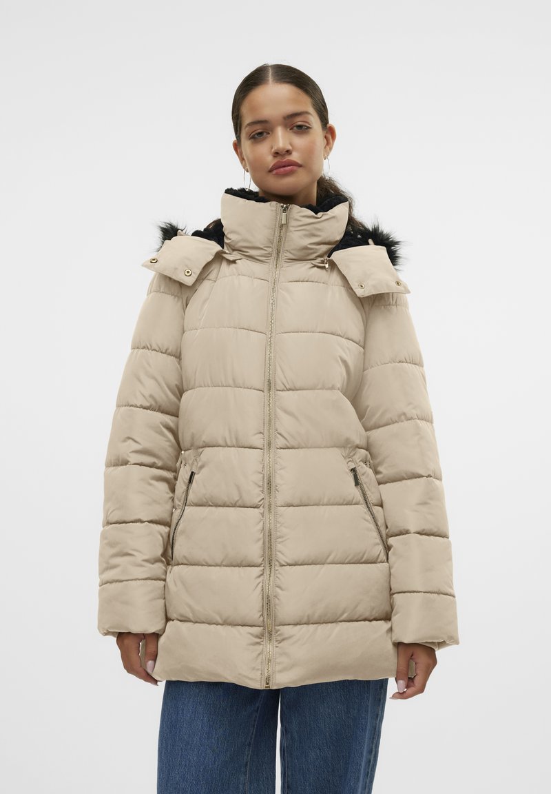 Veste en duvet beige matelassée avec col montant, fermeture éclair, poches avant et accents en fausse fourrure noire sur la capuche. Matériau doux et isolé.