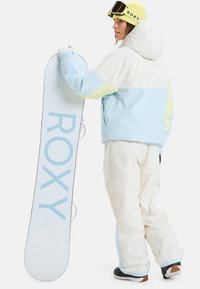 Women's snowboarding outfit featuring a light blue and white jacket with yellow accents, white pants, and a white snowboard labeled "ROXY."