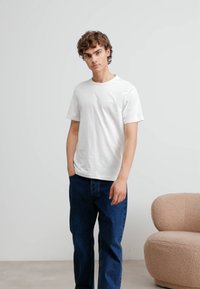 White cotton t-shirt with a crew neck and short sleeves, paired with loose blue denim jeans. Background includes a beige textured armchair.