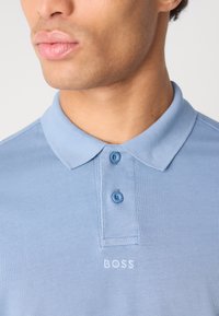 Polo de culoare albastru deschis, realizat dintr-un material moale, având un guler clasic, trei nasturi și un logo brodat "BOSS" pe piept.