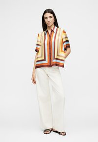 Γυναίκα που φοράει πολύχρωμο ριγέ πουκάμισο με κουμπιά, λευκό παντελόνι με φαρδιά μπατζάκια και μαύρα σανδάλια, στέκεται με το ένα χέρι στην τσέπη.