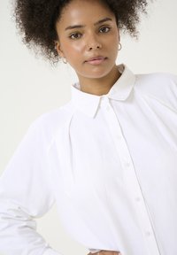 Chemise blanche à boutons avec un col classique, des manches longues et des détails plissés. Fabriquée en tissu léger et lisse.