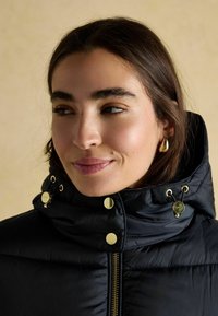 Mladá žena s tmavými vlasmi má na sebe čiernu vypchávkovanú bundu zapnutú po krk so zlatými gombíkmi a zlaté kruhové náušnice, pozerá sa do strany.