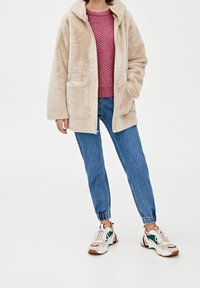 Beige, fluffy fleecejakke med hætte og to forlommer, båret over en lyserød striktrøje, parret med blå jeans og multikolore sneakers.