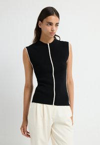 Giovane donna che indossa un top di maglia nera senza maniche con rifiniture bianche al centro e pantaloni bianchi larghi su uno sfondo bianco.