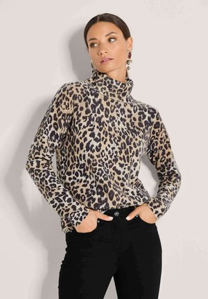 Frau trägt einen hochgeschlossenen Pullover mit Leopardenmuster und schwarze Hose, posiert mit den Händen in den Taschen vor neutralem Hintergrund.